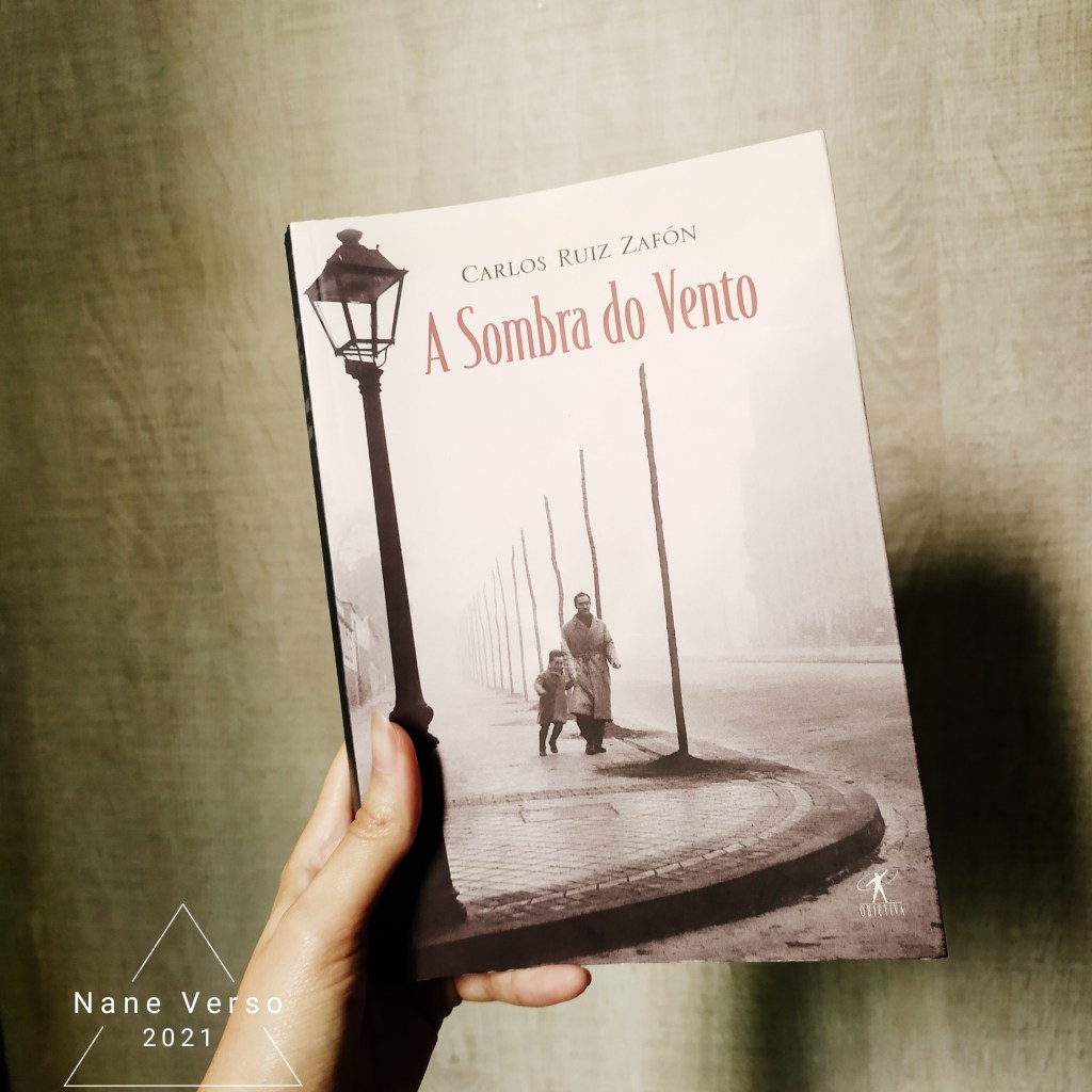 Uma mão segura o livro A sombra do vento em frente a um fundo bege. A foto está com algumas sombras. No canto esquerdo, a marca d'água do blog/instagram: Nane Verso 2021 dentro de um triângulo.